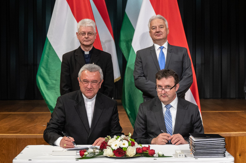 Veres András győri megyéspüspök, az MKPK elnöke és Palkovics László innovációs és technológiai miniszter aláírja a dokumentumot. Mögöttük Semjén Zsolt miniszterelnök-helyettes és Kuminetz Géza, a Pázmány Péter Katolikus Egyetem rektora.